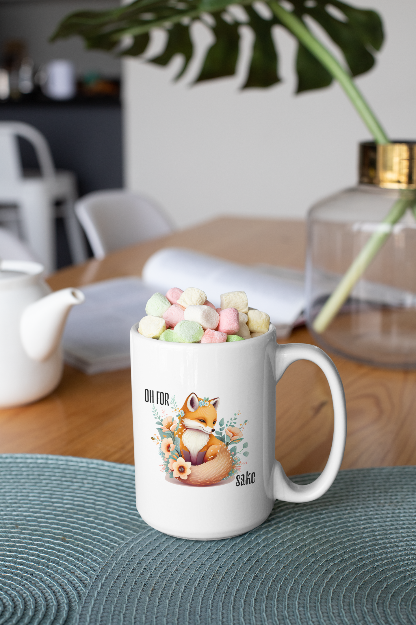 White Oh For Fox Sake Sarcastic Cute Mug on a table with a white tea kettle behind and a large frond in a vase, the mug has pastel colored small marshmallows on top, 15oz with fox with a bushy tail and peach flowers around it