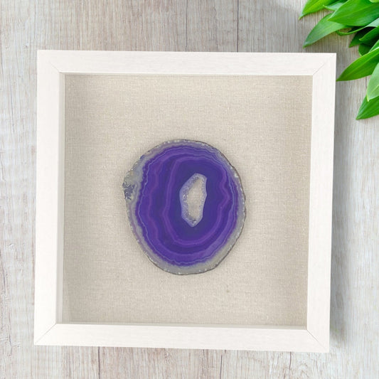 Framed purple agate slice on a light wooden surface with green leaves.