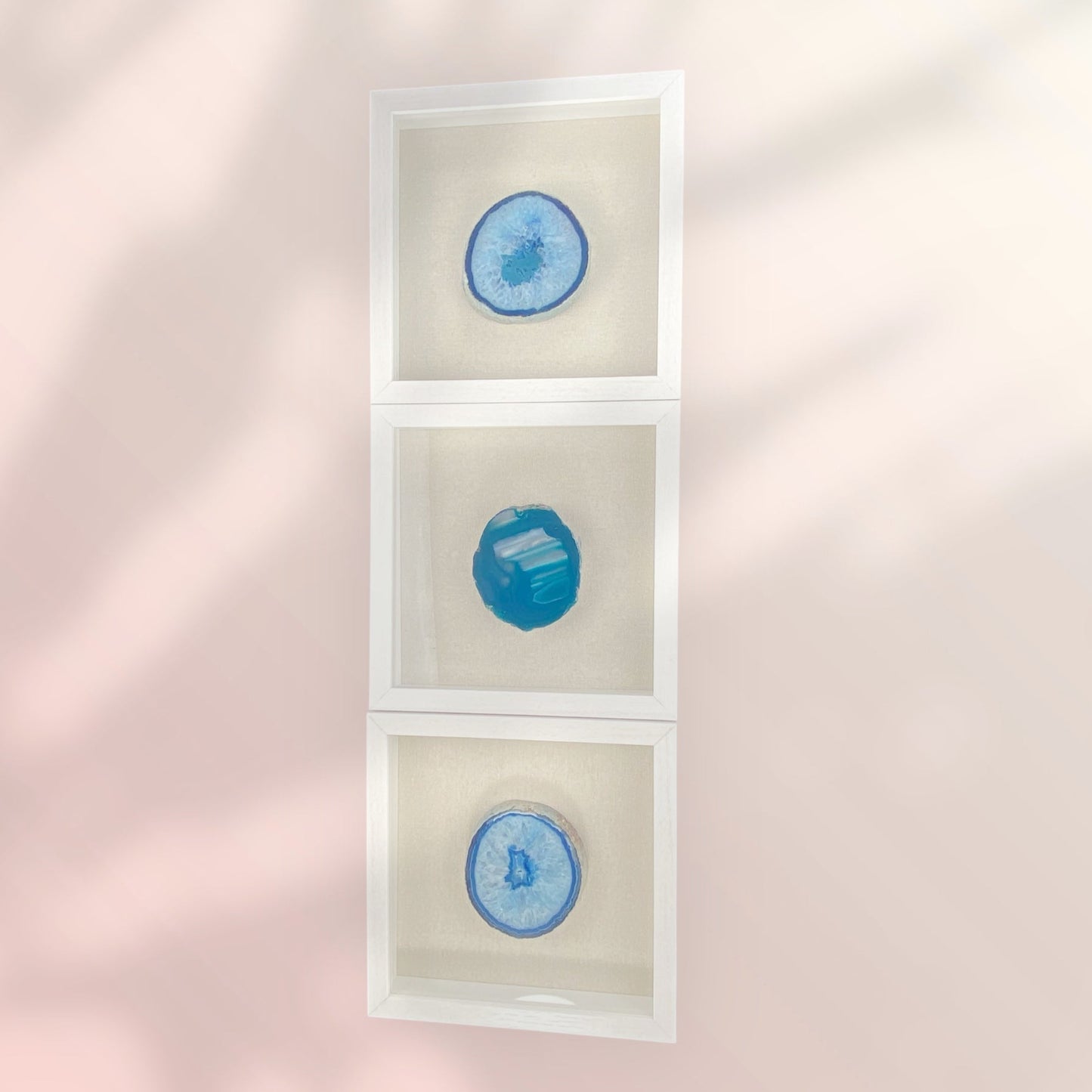 3 stacked white frames with linen backgrounds with one thick blue druzy agate in the center of each frame oriented vertically on a pink wall with shadows