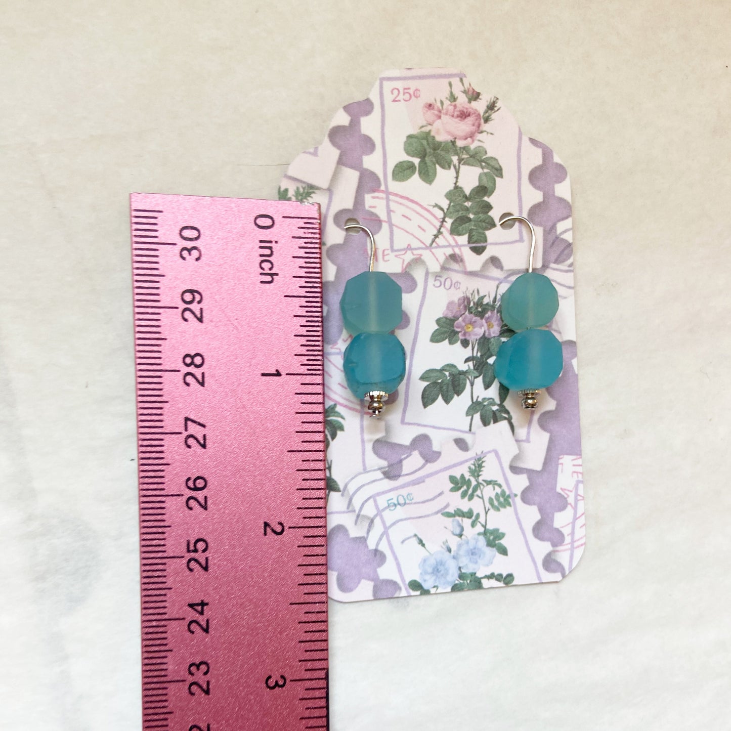 Pair of earrings with blue beads on a floral card next to a ruler for size reference.