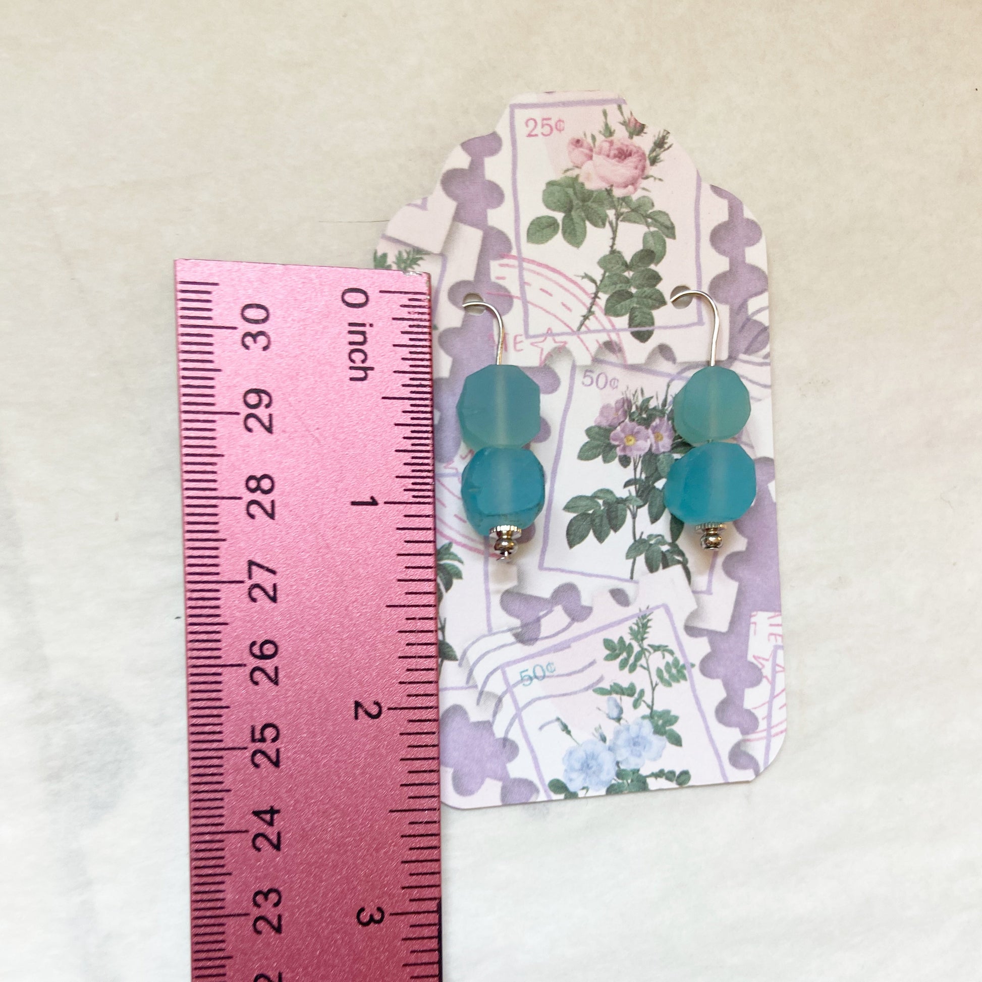 Pair of earrings with blue beads on a floral card next to a ruler for size reference.