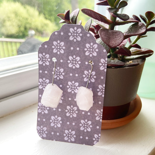 Pair of white earrings on a purple floral earring card with a plant in the background.