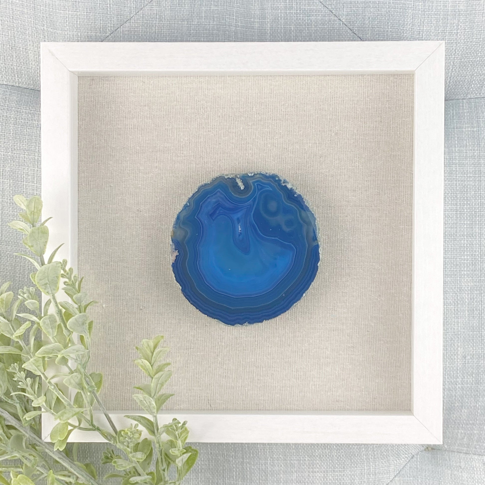 Blue agate stone in a white frame on a textured gray background with green leaves.