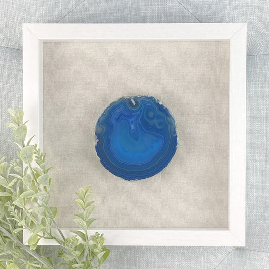 Blue agate stone in a white frame on a textured gray background with green leaves.